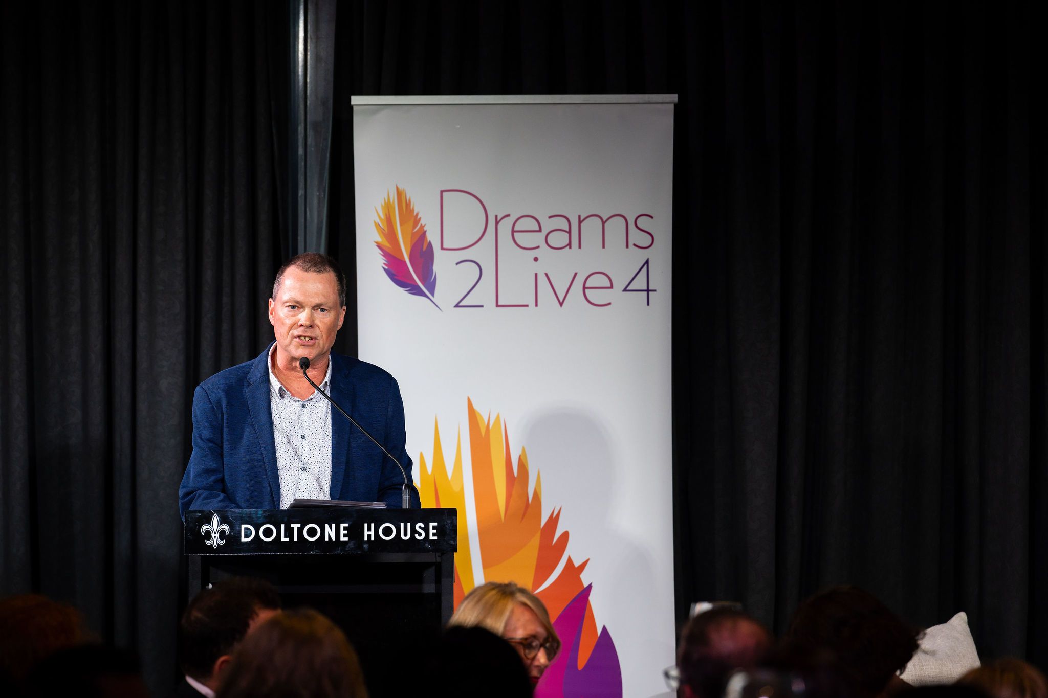 Man at microphone with Dreams2Live4 banner in background